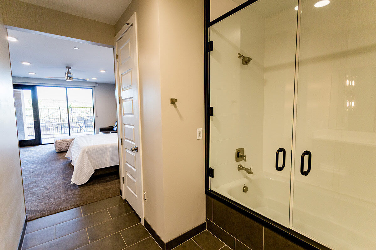 View of shower with glass doors and hotel bedroom through hallway
